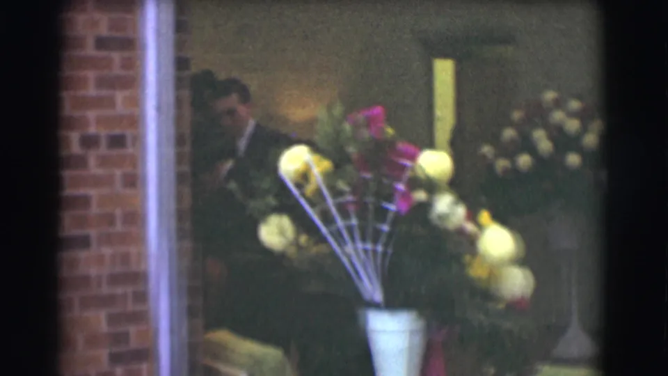 1959 Saint Louis interior frame with a man in a dark suit partly visible behind a large vase of yellow and pink flowers, with brickwork and other floral displays nearby.