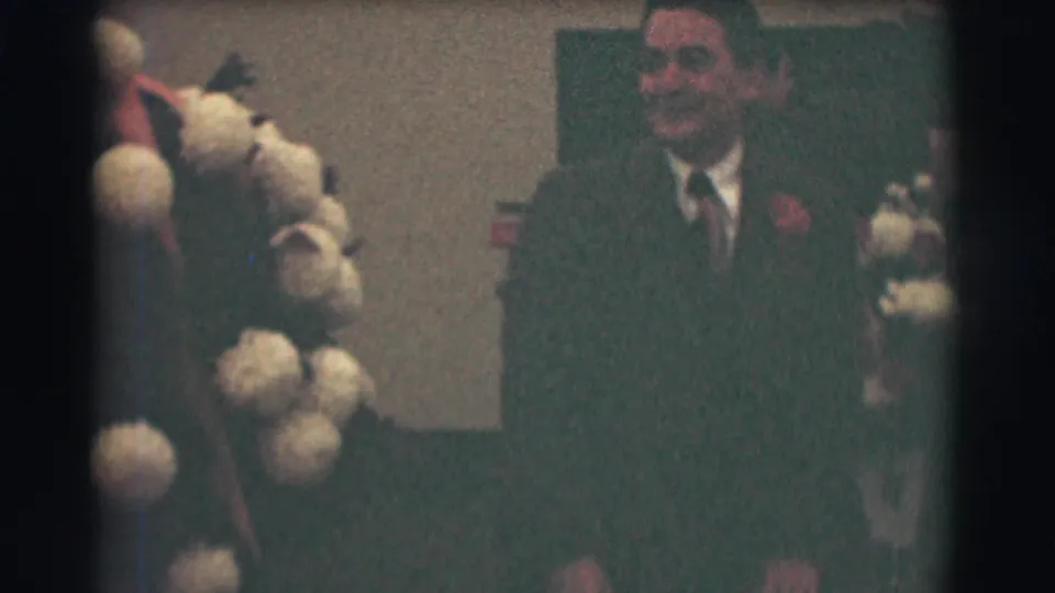 A suited man with a visible face stands near pale pom-pom flowers in a 1959 Saint Louis formal-event home-movie frame.