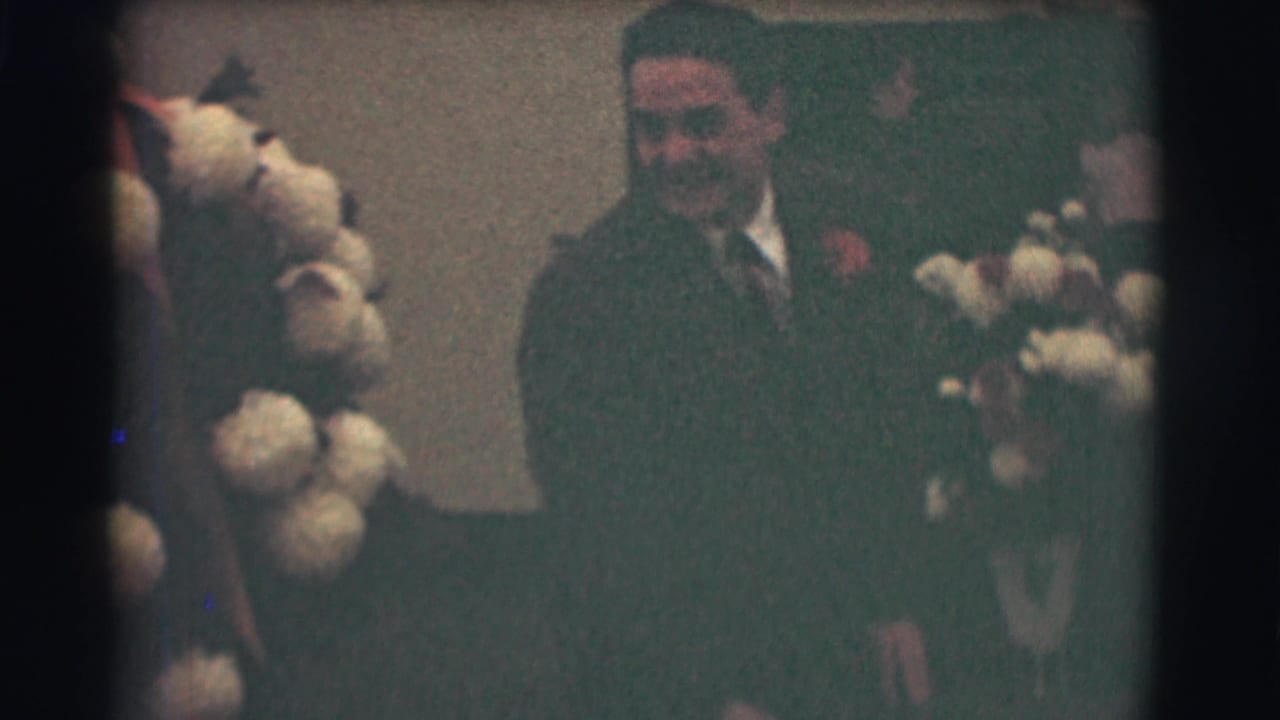 Suited man seated near a tall floral arrangement in a dim indoor room.