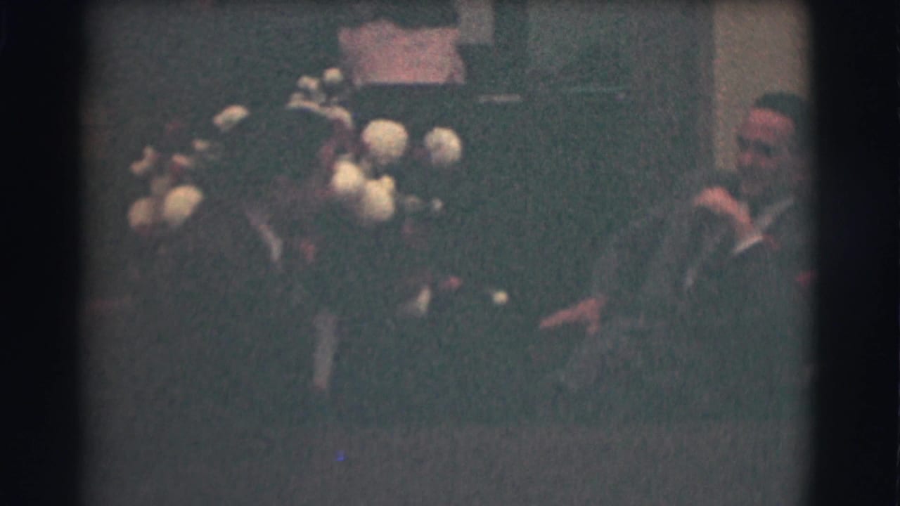 1959 Saint Louis frame with two men in suits seated in conversation near a large floral arrangement in an indoor room.