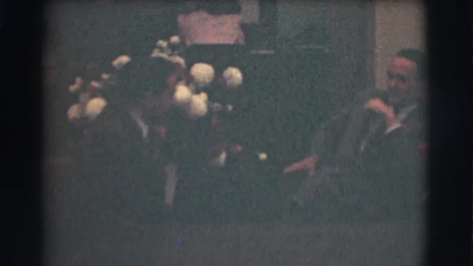 1959 Saint Louis frame with two men in suits seated in conversation near a large floral arrangement in an indoor room.