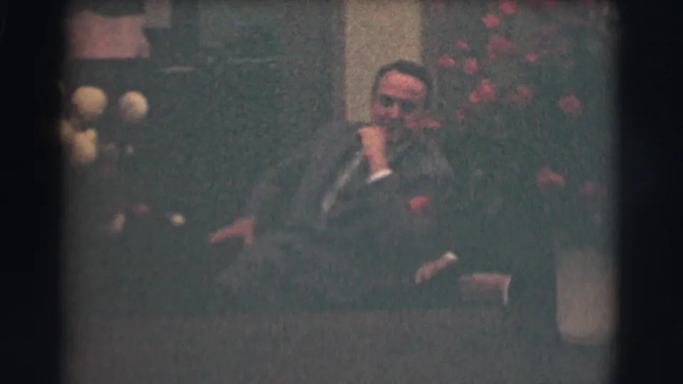 1959 Saint Louis home-movie frame showing a suited man seated beside white and red flowers, with his hand near his face.