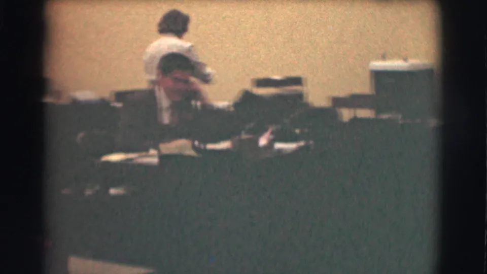 In a 1959 Saint Louis office, a woman stands near the back while a man in a dark suit sits at a desk with papers.