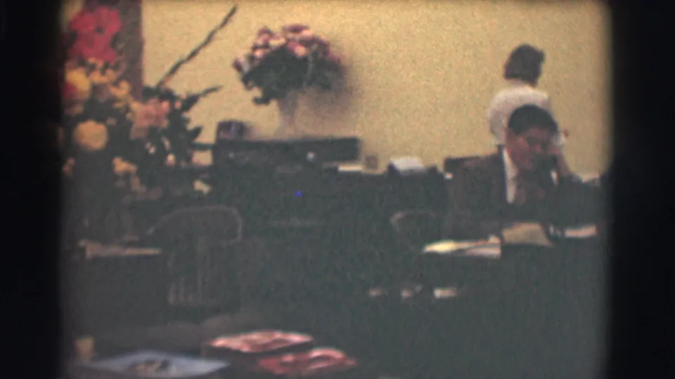1959 Saint Louis office view with a suited man at a desk, a woman standing behind, floral arrangements, and scattered papers.