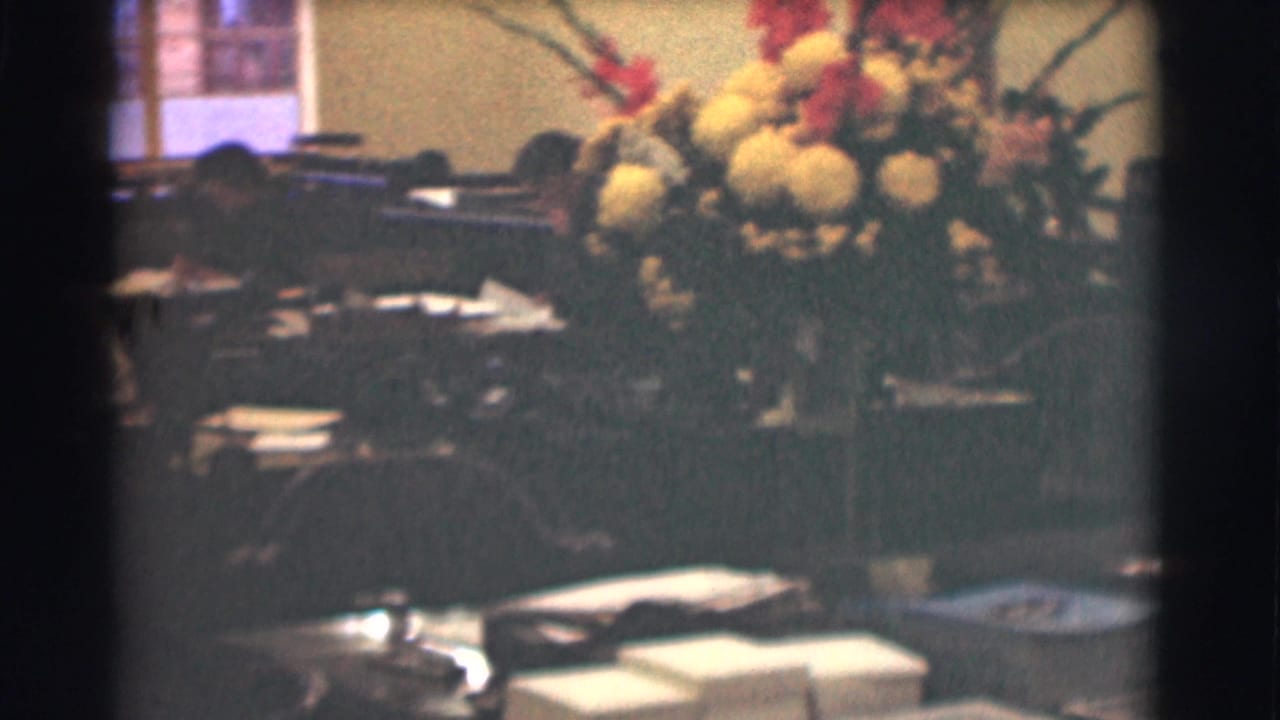 The office view shows a large bouquet beside desks, scattered papers, chairs, and a window in the background.