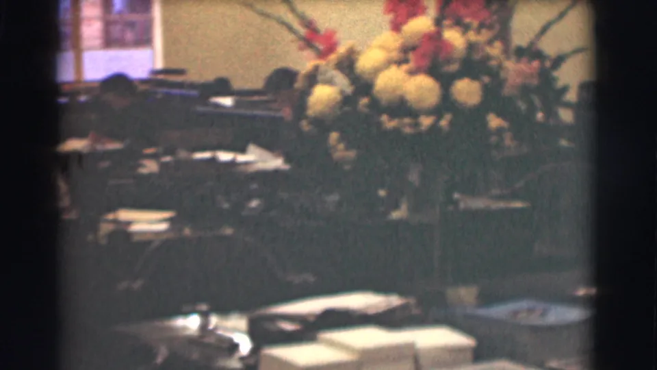 The office view shows a large bouquet beside desks, scattered papers, chairs, and a window in the background.