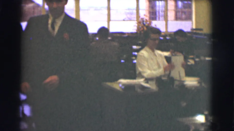 A suited man stands in the foreground while a seated worker handles papers in the Saint Louis office interior.