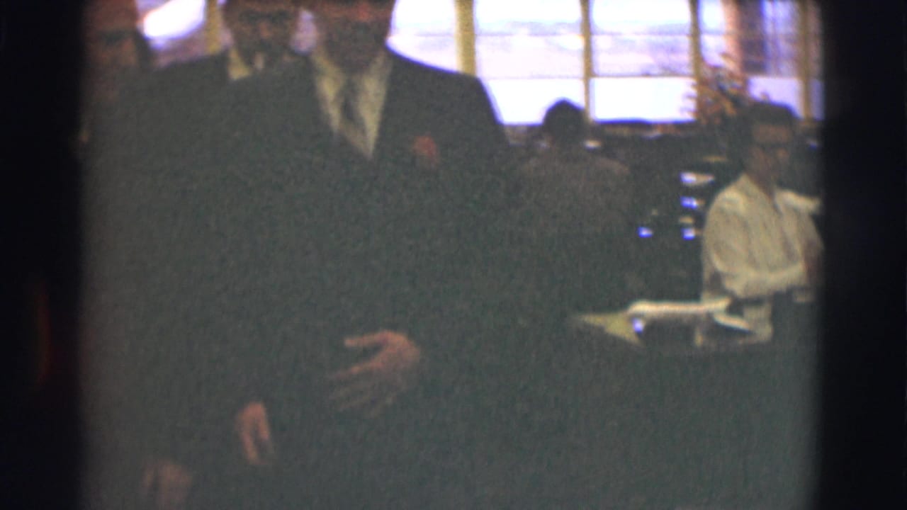 Suited men walking through an office with a seated worker near large windows.