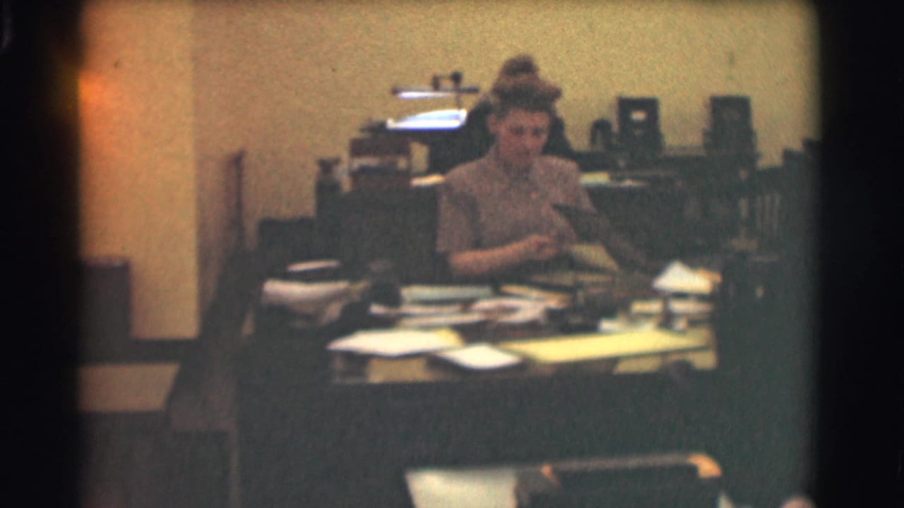 Woman seated at a cluttered office desk with papers and equipment behind her.