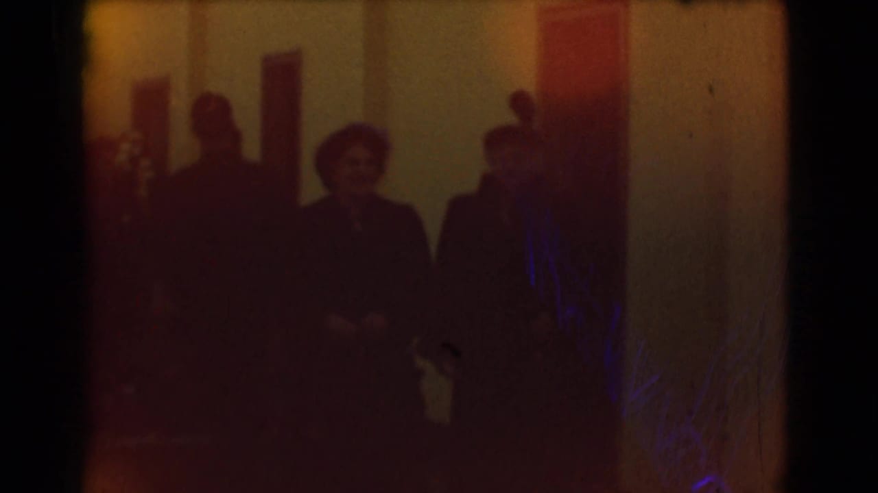 Soft 8mm frame of formally dressed adults in hats moving through an indoor corridor, described as Saint Louis in 1959.