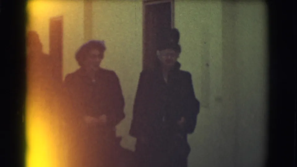 Two formally dressed women, one in a feathered hat, walk with a suited man in a Saint Louis 1959 home-movie corridor scene.