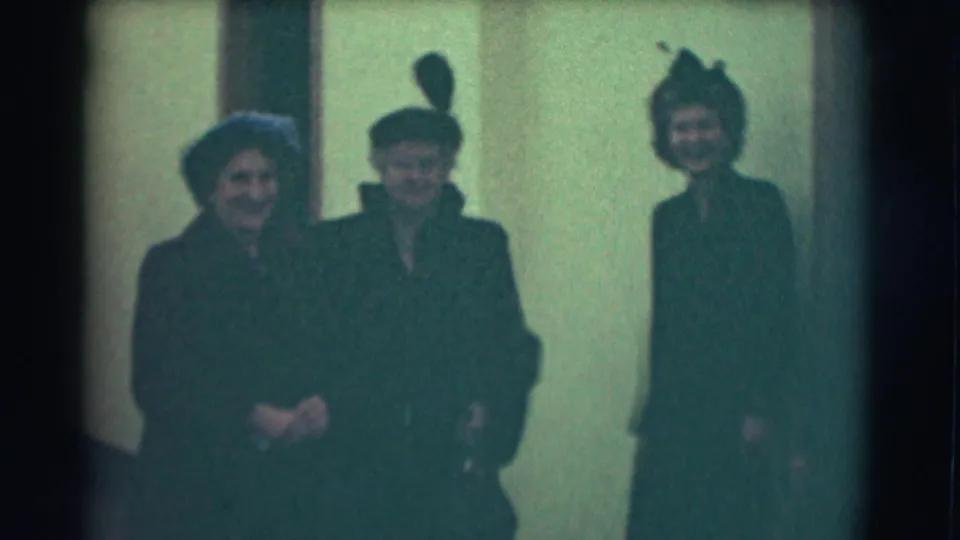 Three formally dressed women in hats appear near a pale interior wall, with one wearing a tall feathered hat in the 1959 home-movie footage.