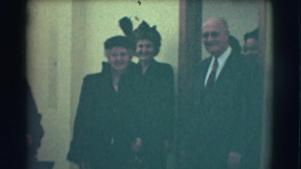 Two women in formal hats and a suited man face the camera in a softly focused 1959 Saint Louis 8mm corridor shot.