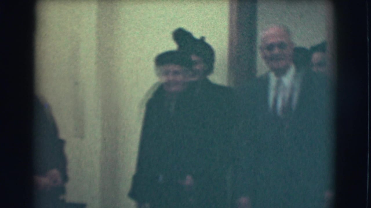 Elderly adults in dark formal clothing stand in a softly lit interior doorway.