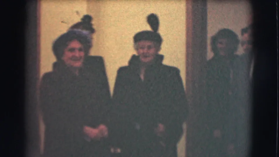 1959 Saint Louis 8mm frame showing two elderly women in formal dark coats and hats standing indoors, with another figure partly visible nearby.