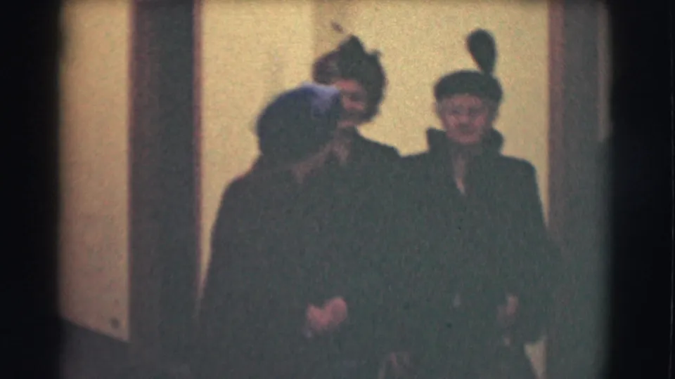 1959 Saint Louis 8mm frame with elderly adults in coats and hats standing close together indoors, some turned away and others facing the camera.