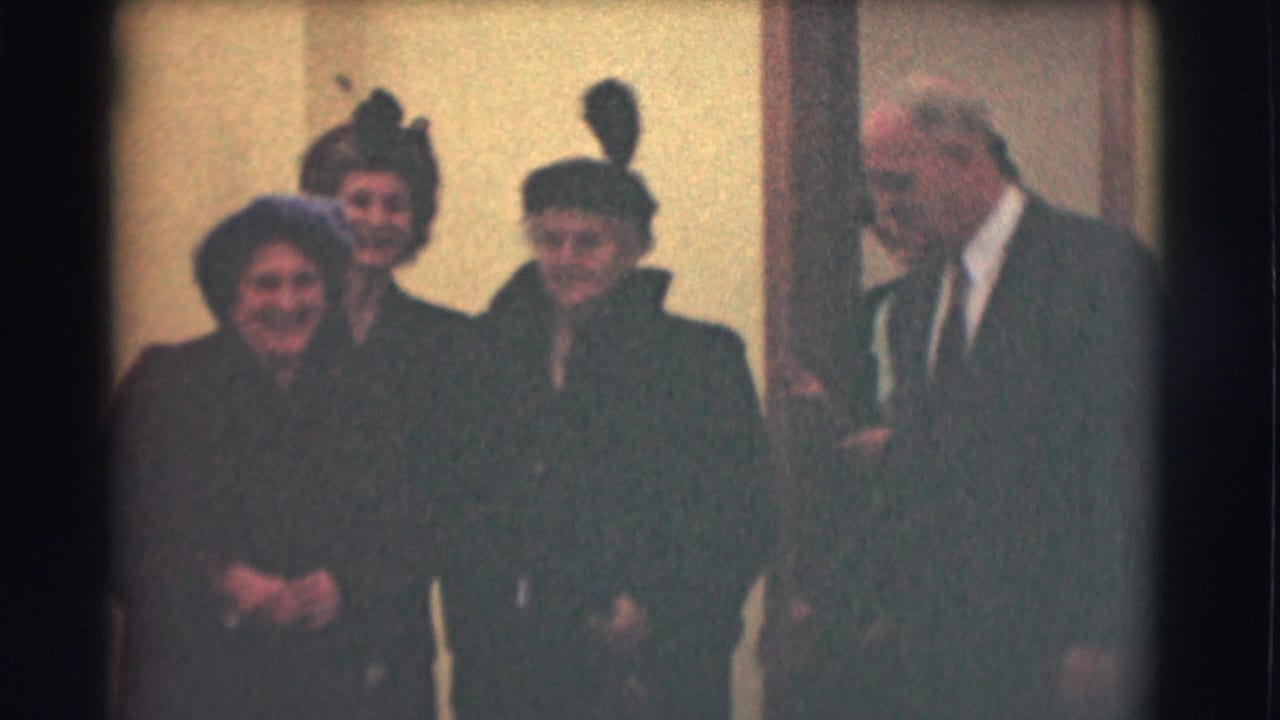 Older women in dark coats and hats stand near a doorway with a suited man beside them.