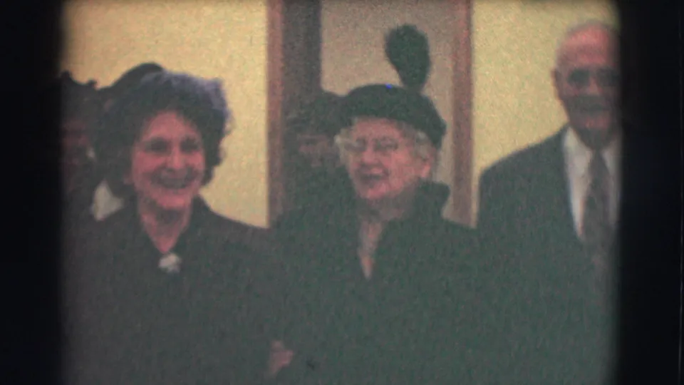 A closer 1959 home-movie view shows two older women in formal hats and dark coats, with a suited man partly visible behind them.