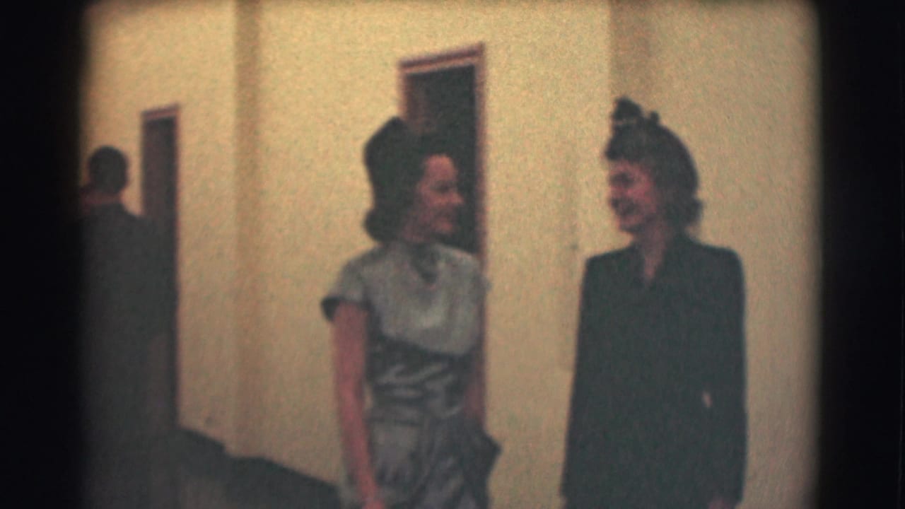 Two women in period dresses and hats stand smiling in an indoor hallway in the Saint Louis 1959 home movie.