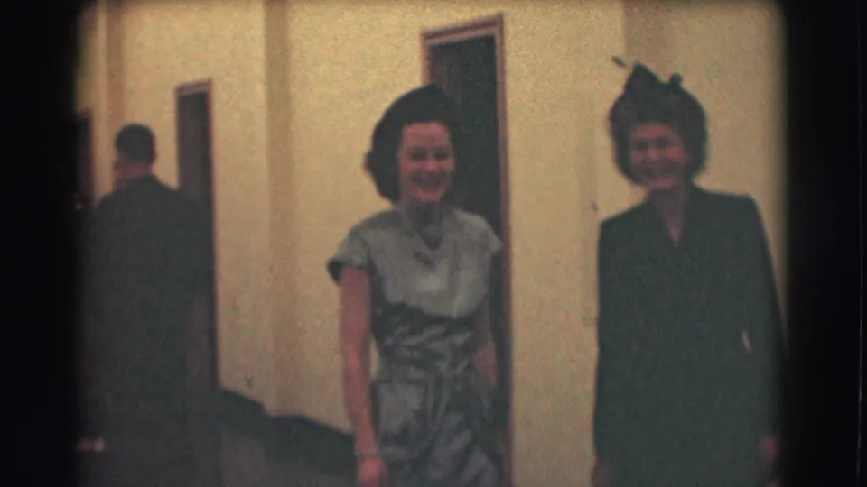 Two women smile side by side in a plain hallway, with another adult partly visible at left.