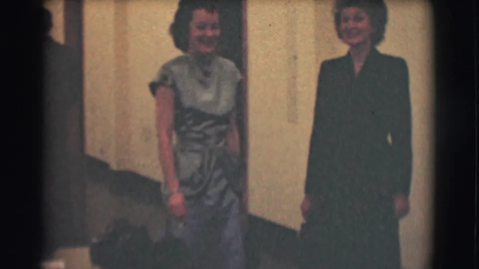 Two women in formal mid-century clothing smile in a Saint Louis hallway, with soft 8mm grain and faded color.