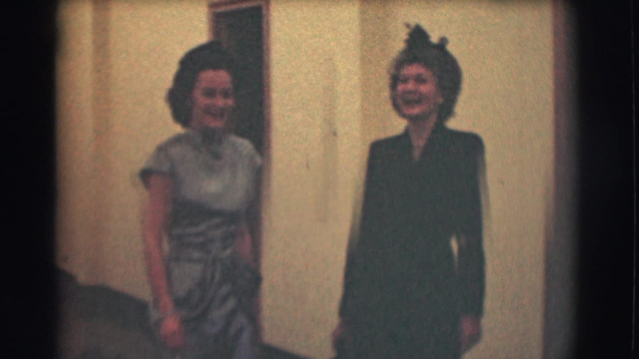Two dressed women stand smiling in a hallway near cream walls and doorways.