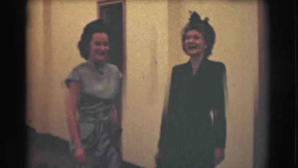 In supported 1959 Saint Louis home-movie footage, two adult women in formal attire stand smiling in a hallway.