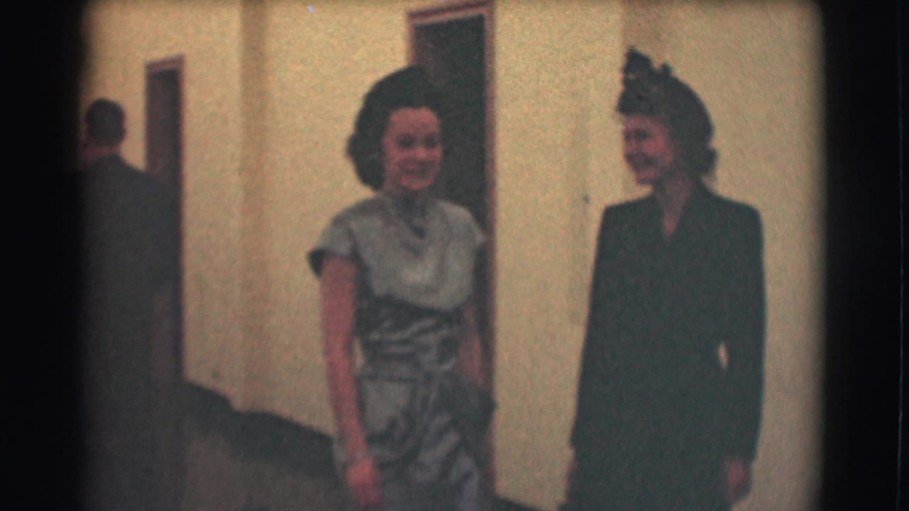 Two formally dressed women appear to converse in a Saint Louis hallway, with a man partly visible in the background.