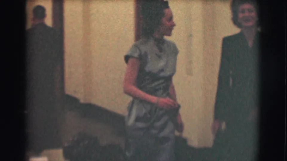 A woman in a fitted light dress turns in profile toward a second woman during the brief hallway exchange.
