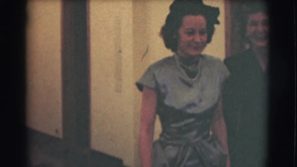 The camera moves closer to the woman in the light dress, with the second formally dressed woman partly visible beside her.