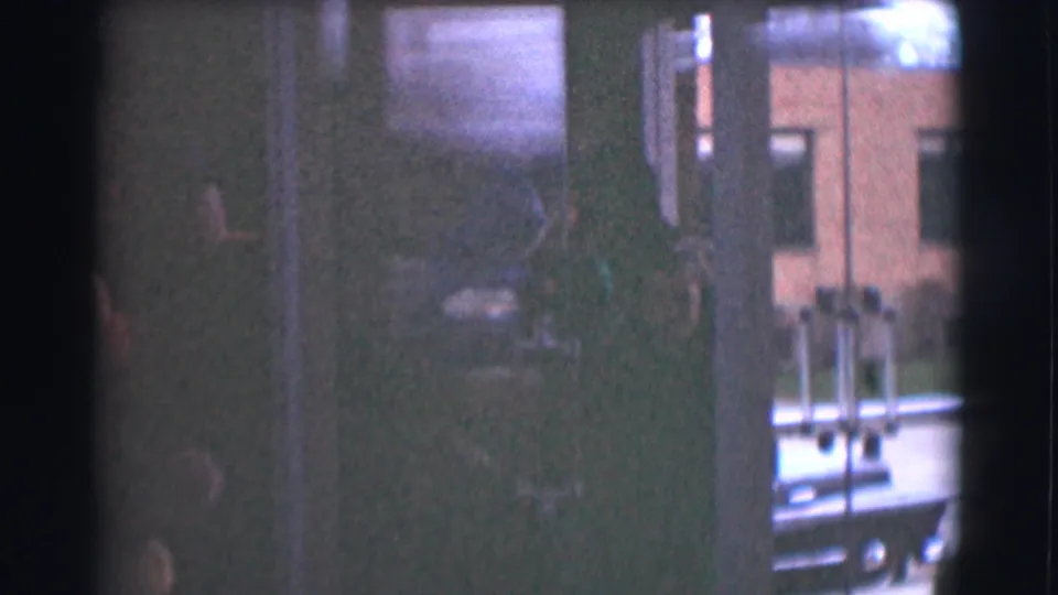The handheld camera shifts toward glass doors or a window, showing dark guest silhouettes and an exterior building beyond the interior.