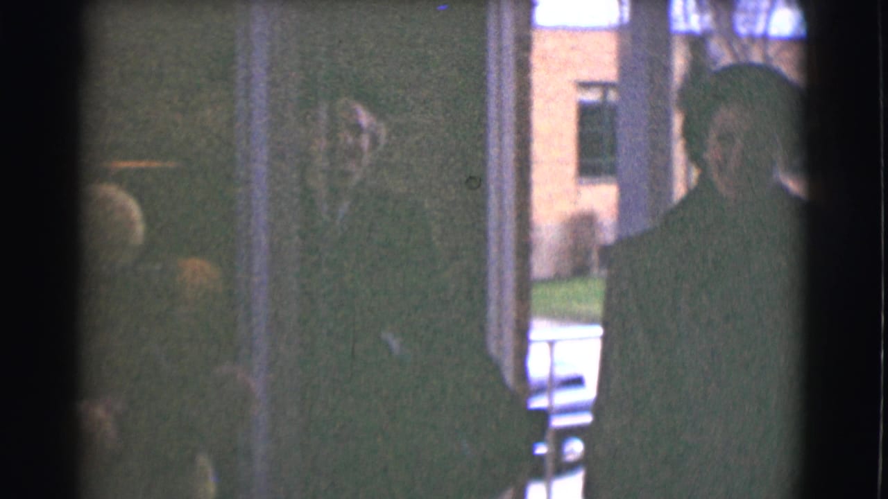 A woman in a dark coat appears near a doorway in Saint Louis, with a second figure at right and brick buildings visible outside.