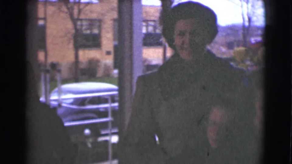 A woman in a dark coat and hat stands near a building entrance, with a parked midcentury car and brick residential buildings behind her.