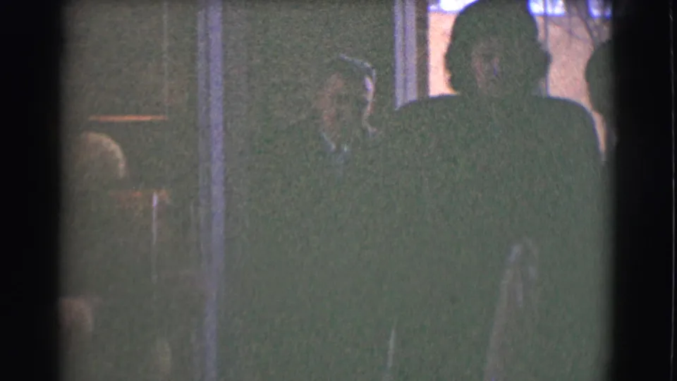 Coated adults stand near an entryway in the 1959 Saint Louis home-movie scene, partly silhouetted against the brighter exterior street.