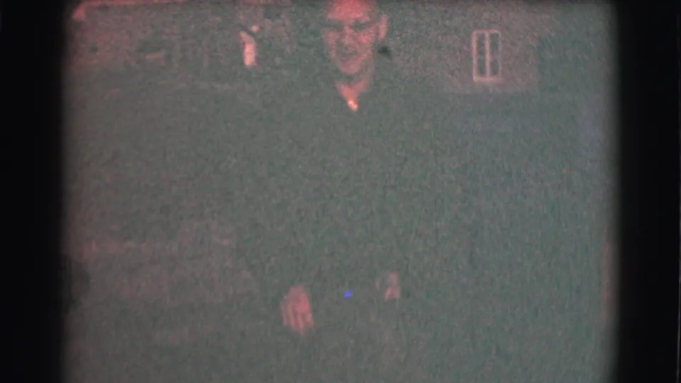Low-exposure 1959 St. Louis home-movie frame with a bundled adult and indistinct residential background forms.
