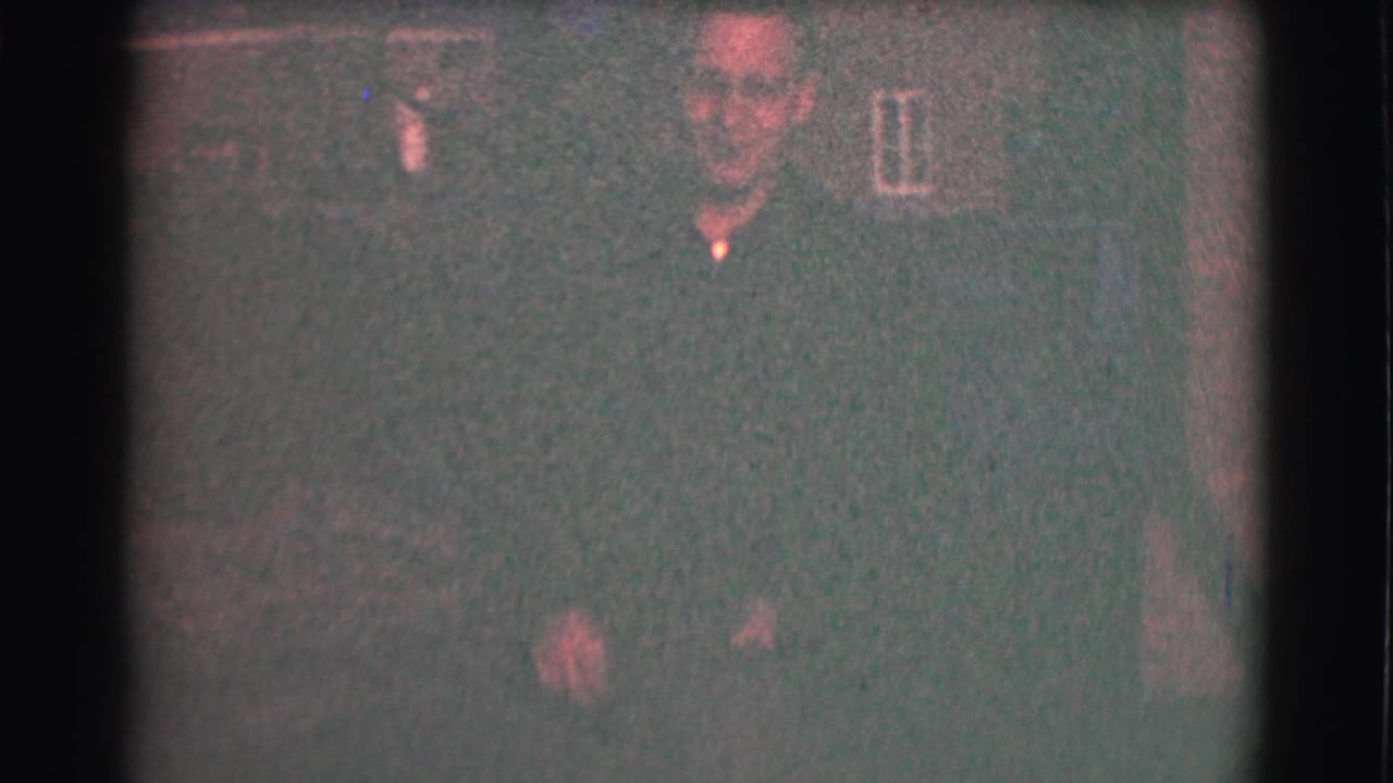 Man in glasses and a dark shirt stands near a brick wall in dim light.