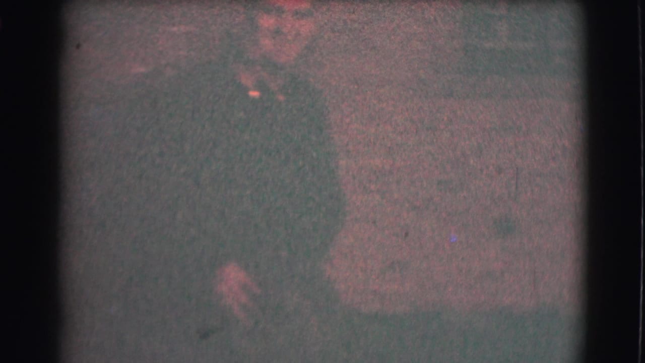 The man in glasses is seen closer against the brick background, with low nighttime exposure and strong film grain.