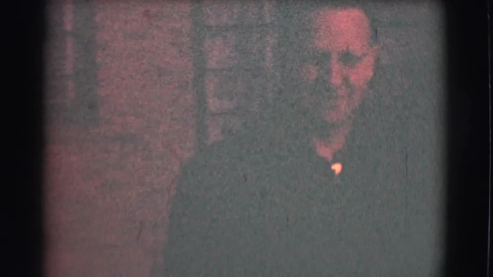 Close, grainy view of the smiling man in glasses and a dark shirt, with the brick wall still visible behind him.