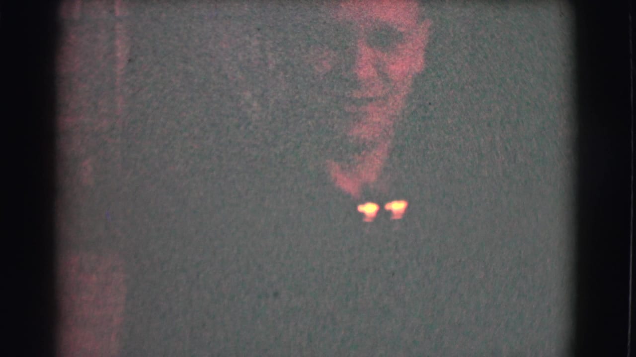 Man with glasses partially visible in a very dark, grainy interior frame.