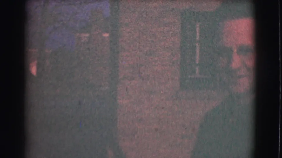 The man with glasses appears at the side of a dim 1959 Saint Louis interior, with framed wall decor and heavy 8mm grain visible.