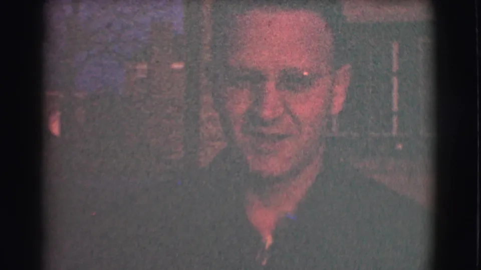 A man with glasses faces the camera in a dark 1959 Saint Louis home-movie frame, with soft focus, reddish cast, and faint background decor.