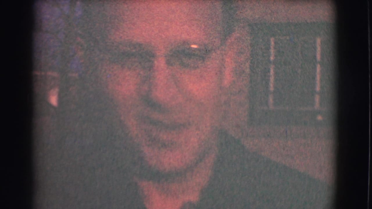 Adult man in glasses smiles toward the camera in a dim close-up.