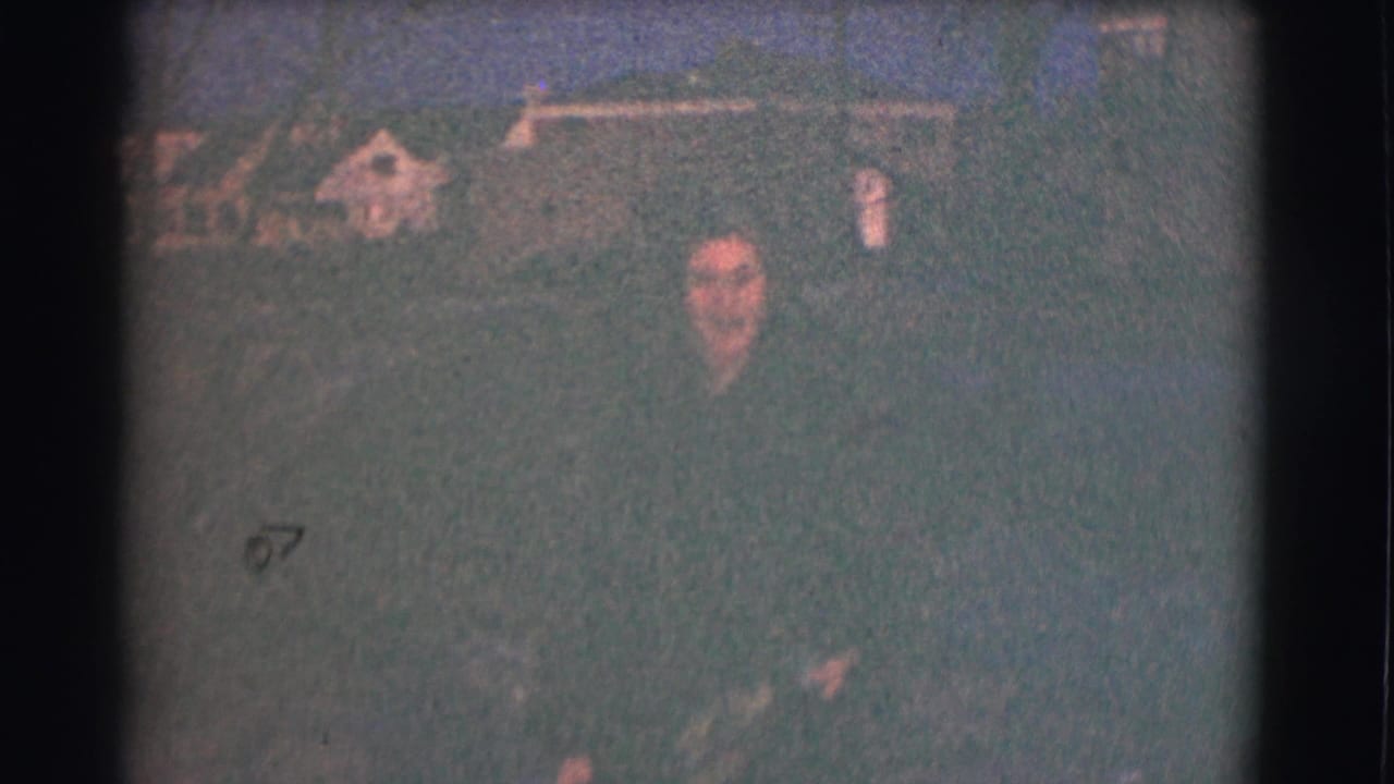 Woman standing outdoors at night in a grainy 8mm frame with faint background shapes.