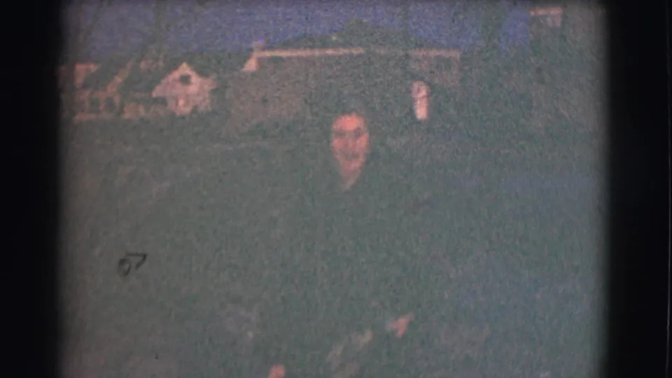 In 1959 Saint Louis home-movie footage, a woman stands outdoors at night while the low-light 8mm image leaves the background soft and indistinct.