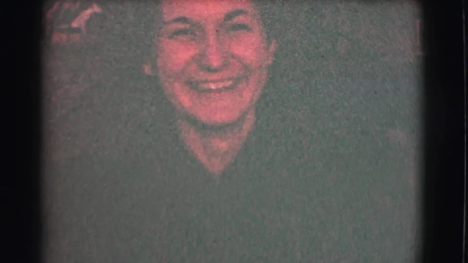 A close 1959 Saint Louis home-movie frame shows the woman smiling broadly, her face lit warmly against a dark, grainy night background.