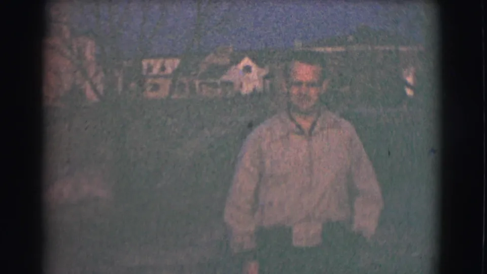 A man stands outdoors in a Saint Louis residential setting, with a house facade and windows visible behind him on grainy 8mm film.