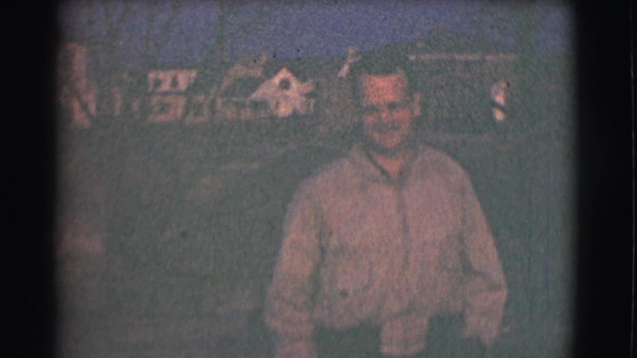 Smiling man in a collared shirt walks toward the camera outdoors with houses in the background.