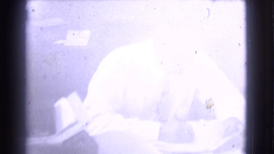 The same adult man appears seated at a desk in a white shirt and tie, writing on papers in the 1959 Saint Louis home movie.
