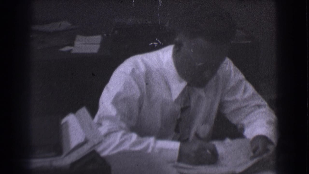 Man in dress shirt and tie writes at a desk in a dim office.