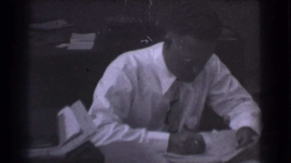 Adult man in glasses, white shirt, and tie writes on paper at a desk in 1959 Saint Louis office footage.
